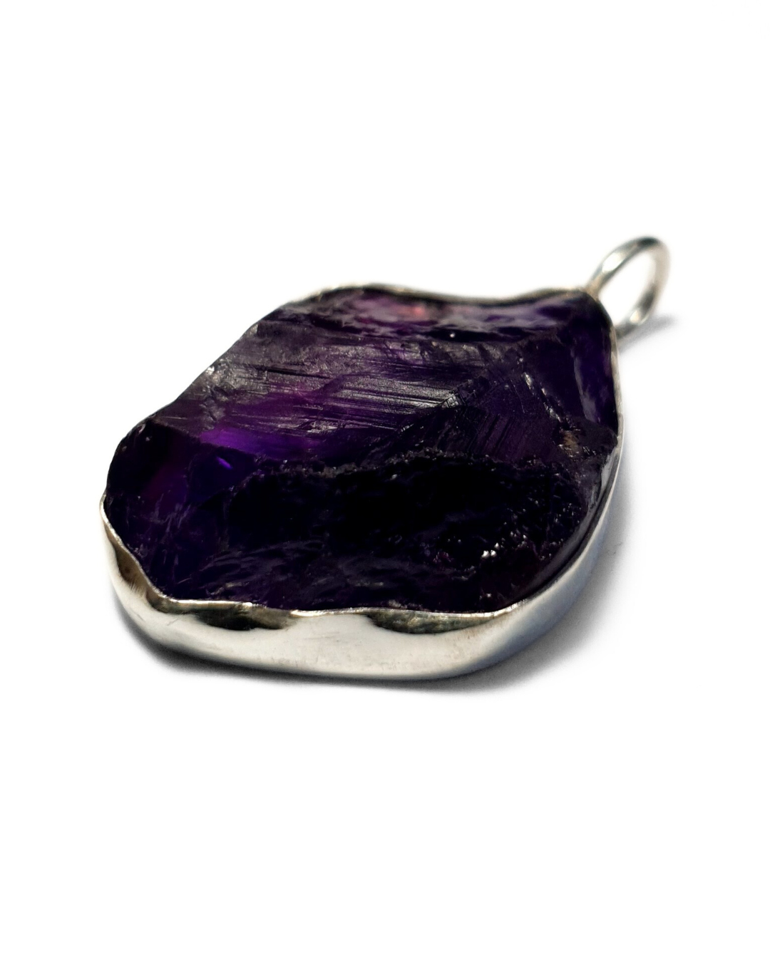 A rough-cut, irregular-shaped amethyst pendant in a silver bezel setting, with a rich and juicy purple color and rough, textured surface. The pendant is on a plain white background