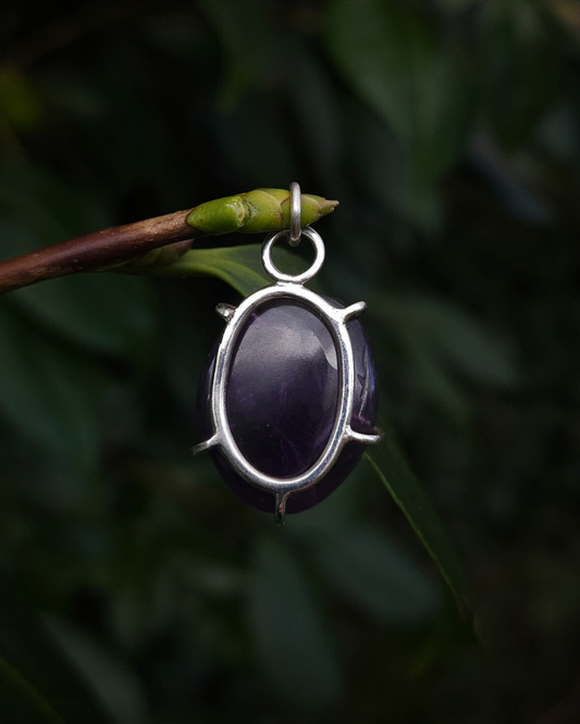 An oval shaped, rough-cut amethyst pendant hangs from a thin budded twig, against a dark green natural background. It is a rich purple colour and is photographed from the back to show how its simple design allows the light to enter the stone and enhance its deep colours and natural textures. The setting is made from recycled sterling silver.