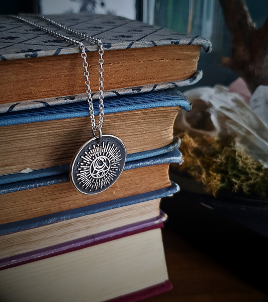A silver pendant with a circular design featuring solar and lunar elements, displayed on a stack of vintage books. The design is the Campfire & Compass Jewellery sigil. It is the solid silver imprint of a wax seal, with the background oxidised a deep black and with the details and edges polished to a bright silver shine in contrast.