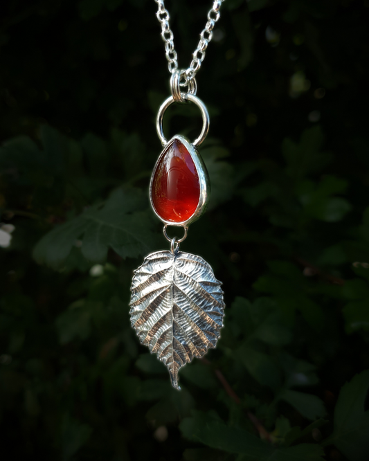 A silver pendant hangs from a chain in front of dark green, out-of-focus background. It is comprised of a smooth, fiery, teardrop-shaped carnelian bezel-set in silver. Below it hangs a solid silver bramble leaf, roughly twice the size of the gemstone, attached by tiny jump rings so it can move freely. The leaf is complete with natural details including veins. The pendant is illuminated in natural sunlight.