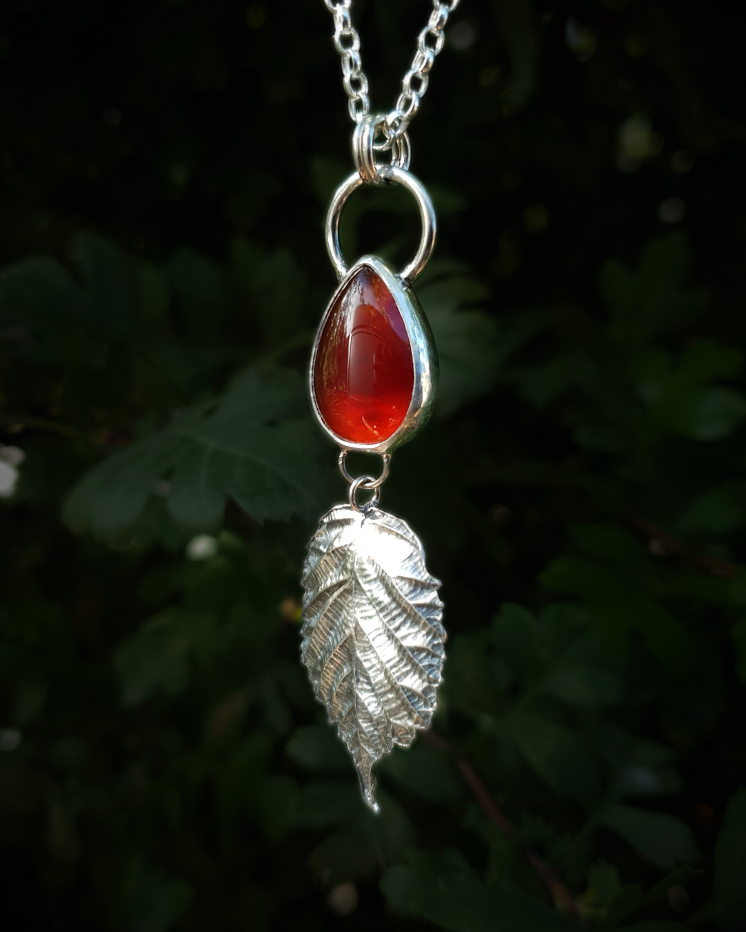 A silver pendant hangs from a chain in front of dark green, out-of-focus background. It is comprised of a smooth, fiery, teardrop-shaped carnelian bezel-set in silver. Below it hangs a solid silver bramble leaf, roughly twice the size of the gemstone, attached by tiny jump rings so it can move freely. The leaf is complete with natural details including veins. The pendant is illuminated in natural sunlight.