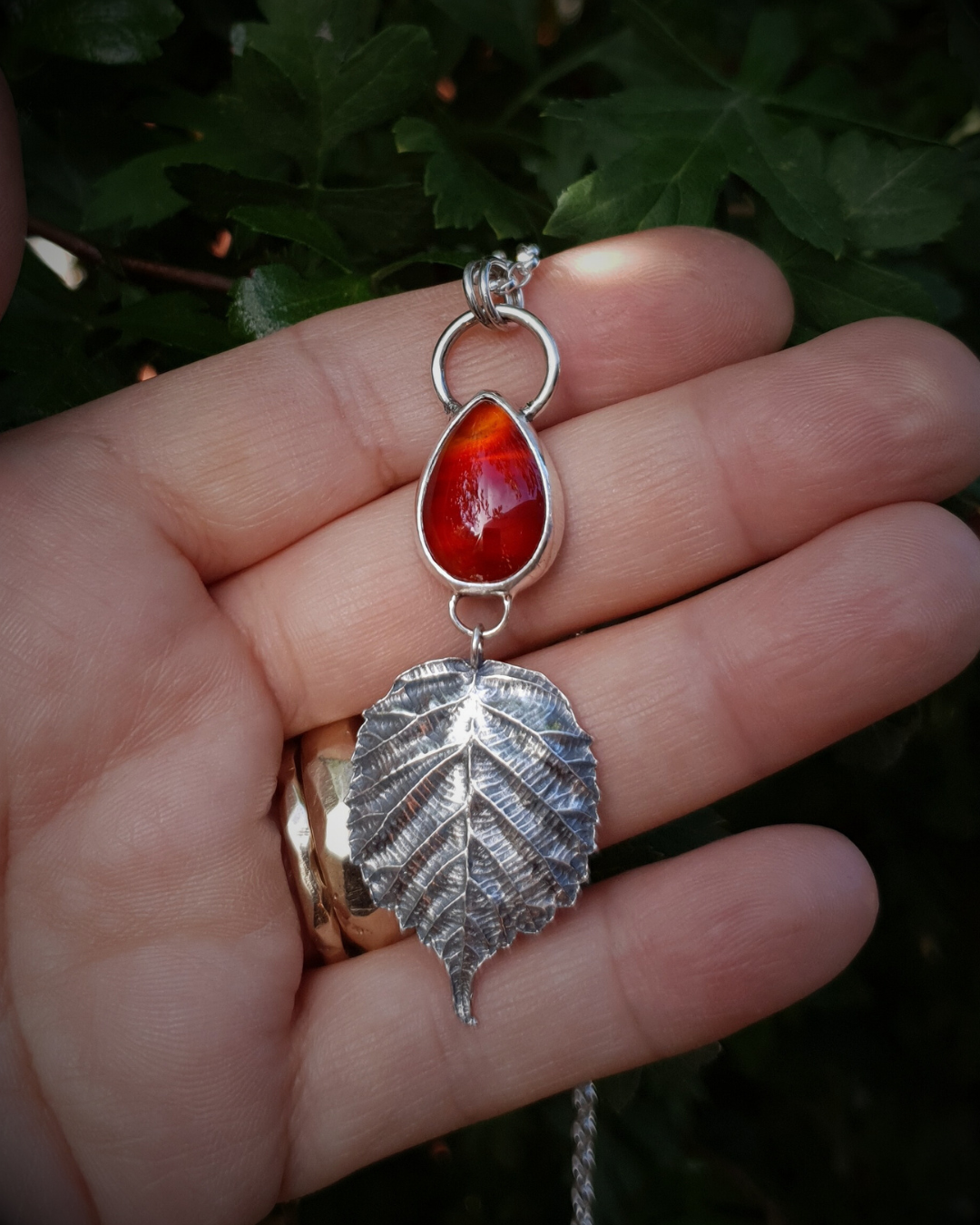 A silver pendant sits in an open palm to demonstrate its size. It is comprised of a smooth, fiery, teardrop-shaped carnelian bezel-set in silver. Below it hangs a solid silver bramble leaf, roughly twice the size of the gemstone, attached by tiny jump rings so it can move freely. The leaf is complete with natural details including veins. The pendant is illuminated in natural sunlight and the carnelian shows beautiful banding of different orange shades.