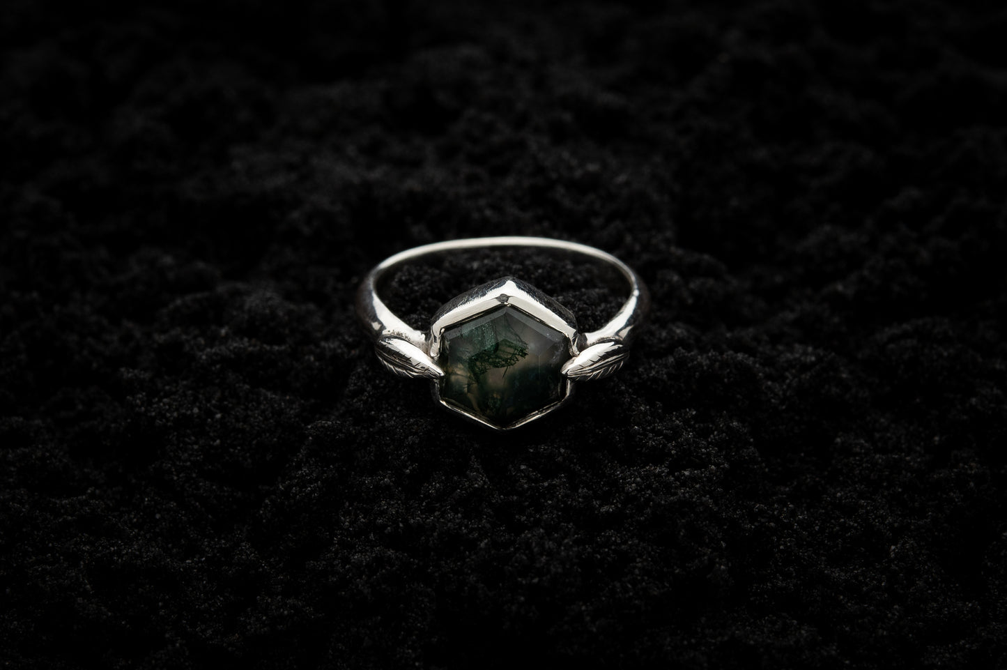 A leafy engagement ring rests on black sand and is lit in soft, glowy light.
A luscious hexagonal moss agate is set in a white gold setting with a slim leaf on either side, folded slightly over the stone.