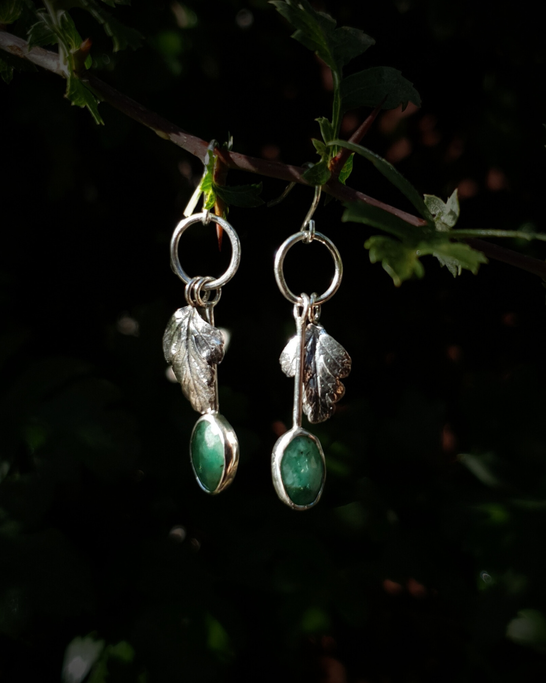 Two silver hook earrings hang from a leafy branch in front of dark green, out-of-focus background. They are comprised of a little silver circlet from which hangs a solid silver leaf. A faceted emerald also hangs from the circlet at the end of a long stem so it hangs below the leaf. The emeralds are rose-cut and rather unrefined showing different shades of green and the leaves are complete with natural details including veins. The earrings are illuminated in natural sunlight.