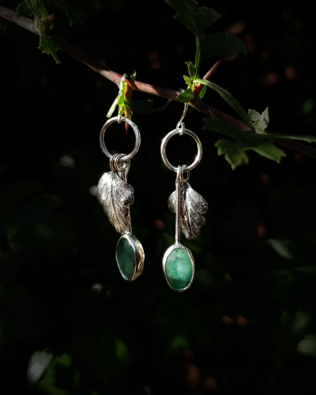 Two silver earrings hang from a leafy branch in front of dark green, out-of-focus background. They are comprised of a little silver circlet from which hangs a solid silver leaf. A faceted emerald also hangs from the circlet at the end of a long stem so it hangs below the leaf. The emeralds are rose-cut and rather unrefined showing different shades of green and the leaves are complete with natural details including veins. The earrings are illuminated in natural sunlight.