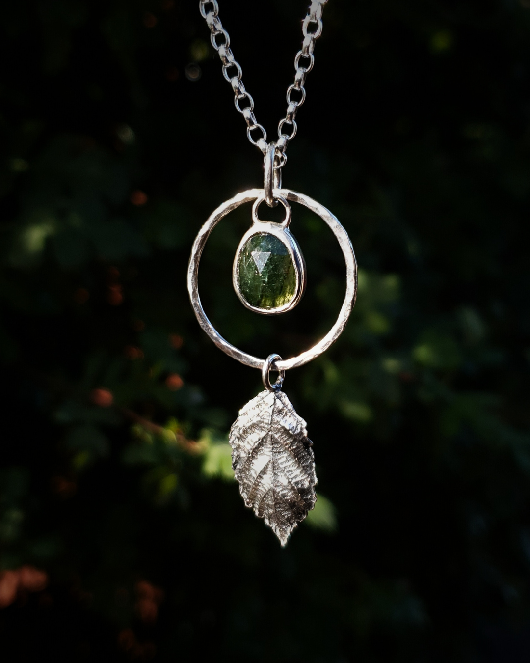 A silver pendant hangs from a chain in front of dark green, out-of-focus background. It is comprised of a rose-cut, green rutile quartz bezel-set in silver and surrounded by a hammered silver halo-like circlet. A solid silver bramble leaf hangs rom the bottom of the circlet. Everything is attached by tiny jump rings so it can move naturally. The leaf is oxidised and polished to reveal the natural details including veins. The pendant is illuminated in natural sunlight.