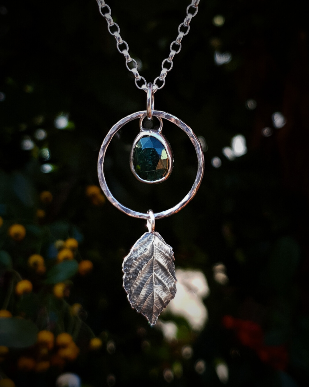 A silver pendant hangs from a chain in front of dark green, out-of-focus background. It is comprised of a rose-cut, green rutile quartz bezel-set in silver and surrounded by a hammered silver halo-like circlet. A solid silver bramble leaf hangs rom the bottom of the circlet. Everything is attached by tiny jump rings so it can move naturally. The leaf is oxidised and polished to reveal the natural details including veins. The pendant is illuminated in natural sunlight.