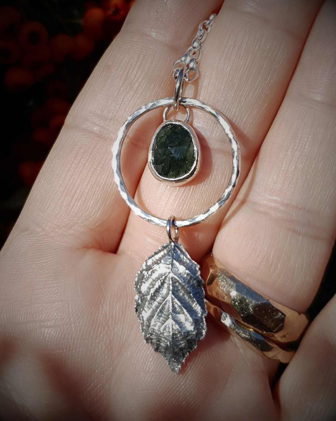 A silver pendant lays on an open palm to demonstrate its size. It is comprised of a rose-cut, green rutile quartz bezel-set in silver and surrounded by a hammered silver halo-like circlet. A solid silver bramble leaf hangs rom the bottom of the circlet. Everything is attached by tiny jump rings so it can move naturally. The leaf is oxidised and polished to reveal the natural details including veins. The pendant is illuminated in natural sunlight.