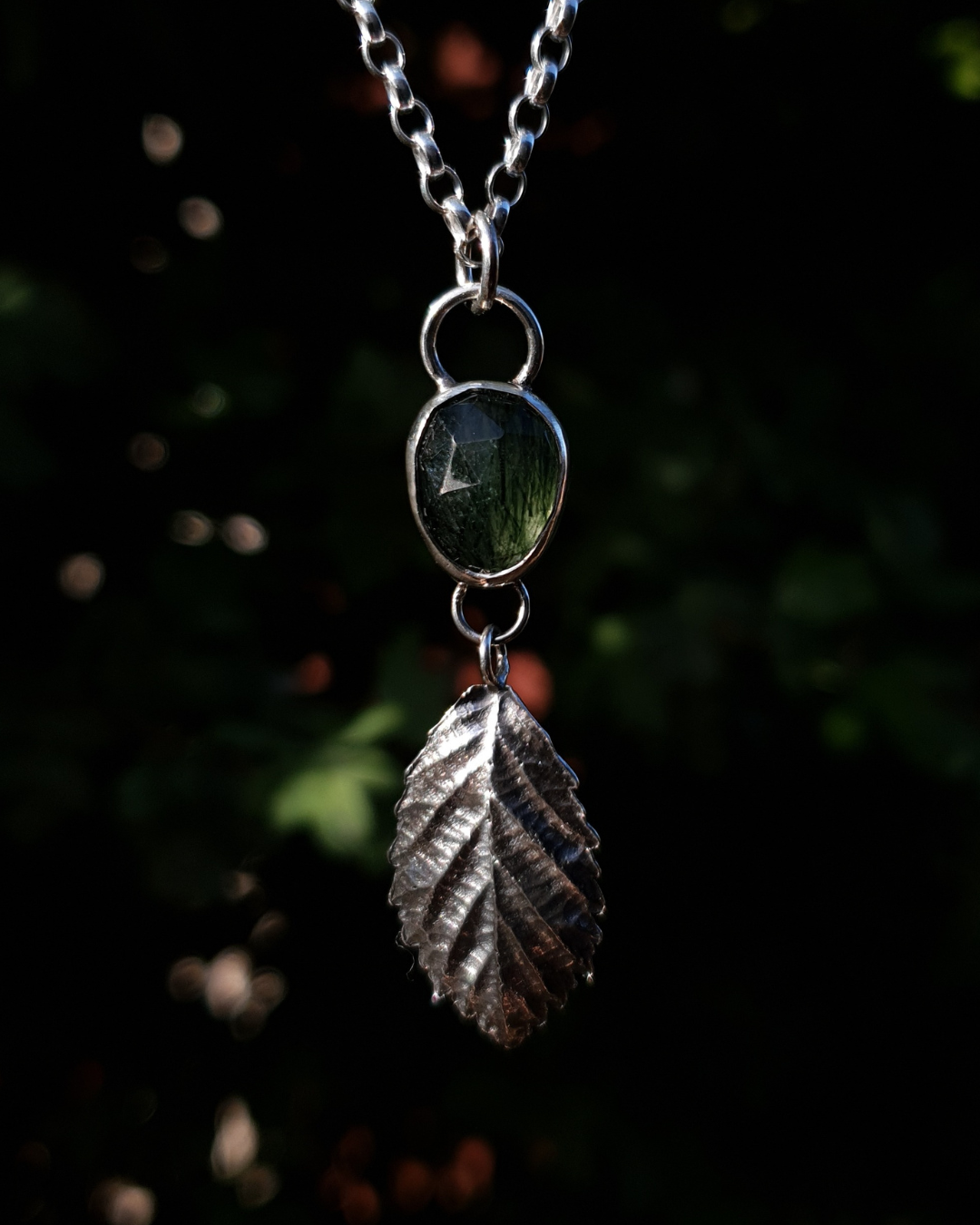 A silver pendant hangs from a chain in front of dark green, out-of-focus background. It is comprised of a rose-cut, green rutile quartz bezel-set in silver. Below it hangs a solid silver bramble leaf, roughly twice the size of the gemstone, attached by tiny jump rings so it can move freely. The leaf is complete with natural details including veins. The pendant is illuminated in natural sunlight.
