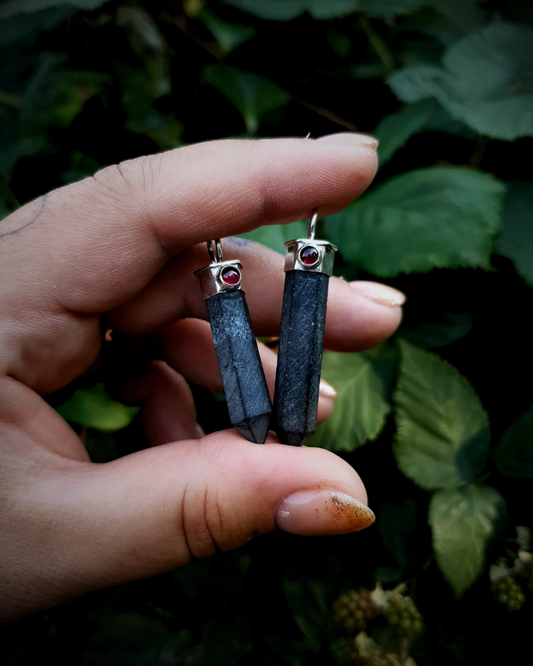 Two black tourmaline point pendants are held between a finger and a thumb. They are set in silver settings with a small round garnet in the front of each one. The background is a dark, slightly out of focus bramble bush