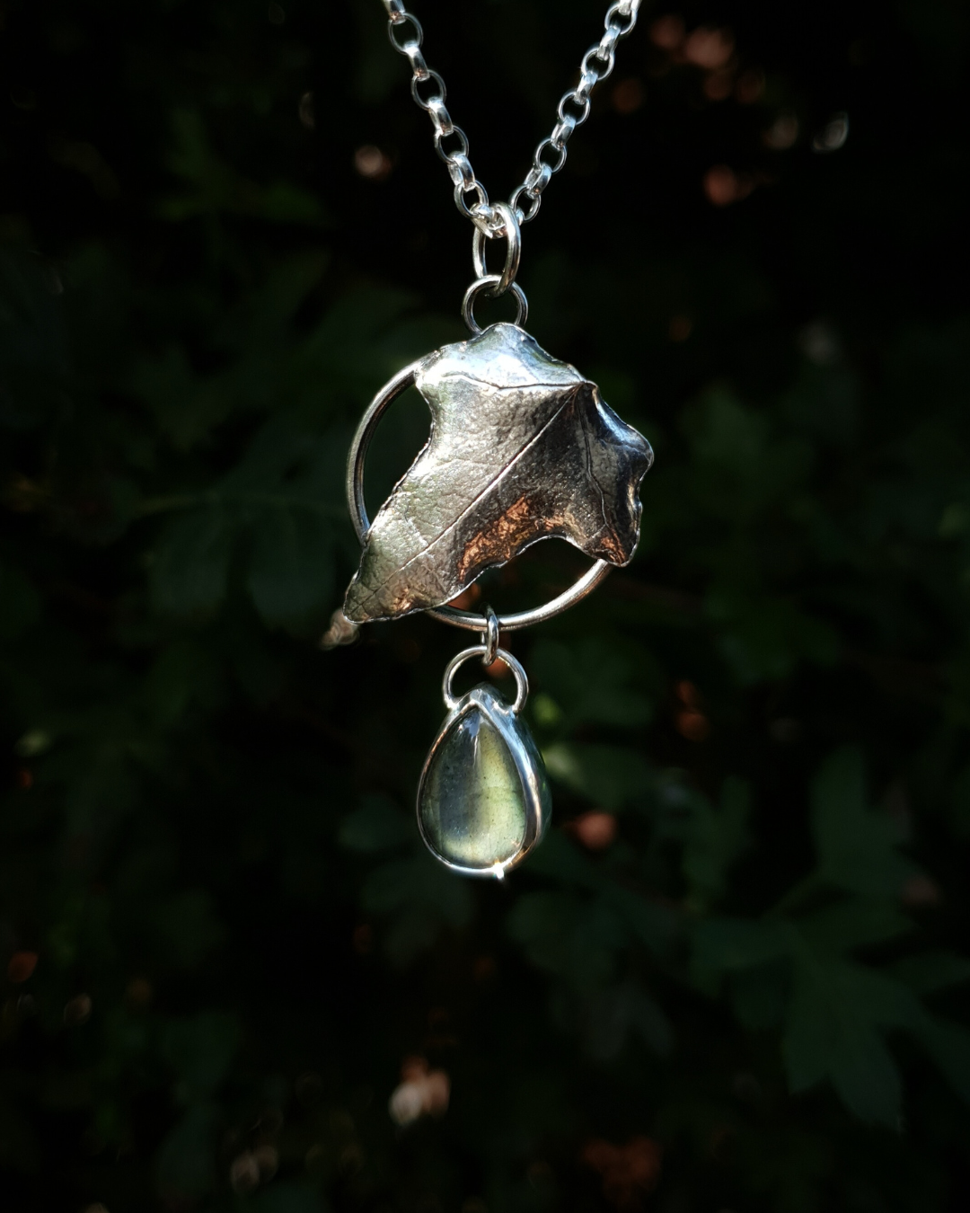Silver pendant comprised of a silver circlet from which a solid silver ivy leaf hangs from a chain. A small teardrop shaped labradorite hangs below it from the circlet. The labradorite is shining dark turquoise and a golden-green in the light; and the ivy leaf is complete with natural details including veins. In the background is out of focus natural greenery and the pendant is bathed in natural sunlight.