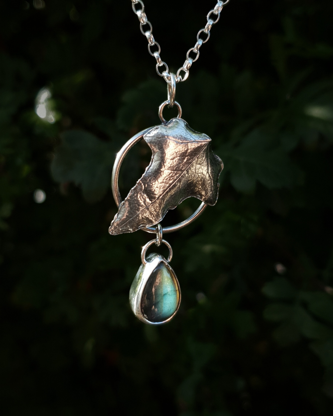 Silver pendant comprised of a silver circlet from which a solid silver ivy leaf hangs from a chain. A small teardrop shaped labradorite hangs below it from the circlet. The labradorite is shining dark blue and a pale irridescent turquoise in the light; and the ivy leaf is complete with natural details including veins. In the background is out of focus natural greenery and the pendant is bathed in natural sunlight.