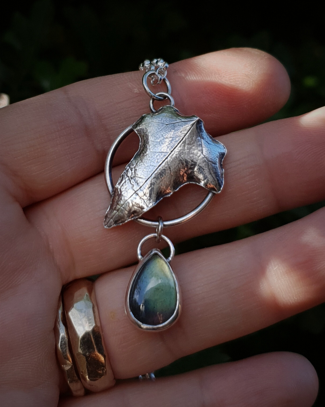 Silver pendant comprised of a silver circlet from which a solid silver ivy leaf hangs from a chain. A small teardrop shaped labradorite hangs below it from the circlet. The labradorite is shining dark blue, light green and a golden-yellow in the light; and the ivy leaf is complete with oxidised natural details including veins. The pendant is held in an open hand, which wears two gold rings, to demonstrate its size. There is scattered sunlight across the hand.