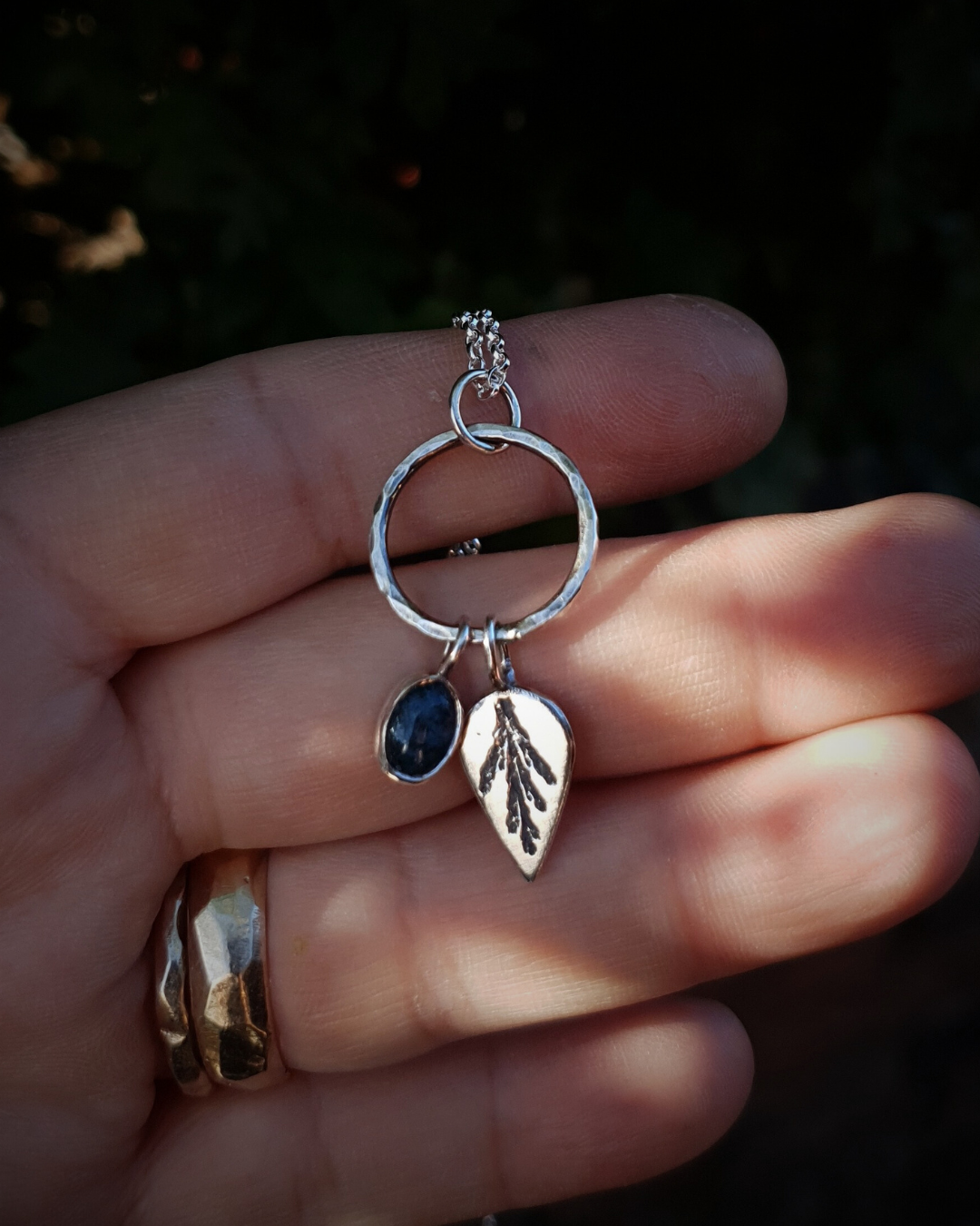 Silver pendant comprised of a silver circlet from which a solid silver upside-down teardrop, imprinted with a cypress sprig, and moss agate pendant hang from a silver chain. The moss agate is a small oval with dark green and white details; and the teardrop charm is approximately twice it's length.
The pendant lays in an open hand, wearing two gold rings, to demonstrate its size.
In the background is out of focus natural greenery.