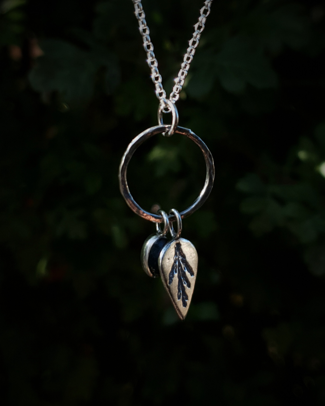 Silver pendant comprised of a silver circlet from which a solid silver upside-down teardrop, imprinted with a cypress sprig, and moss agate pendant hang from a silver chain. The moss agate is a small oval with dark green and white details; and the teardrop charm is approximately twice it's length. In the background is out of focus natural greenery and the charms are illuminated in natural sunlight.
