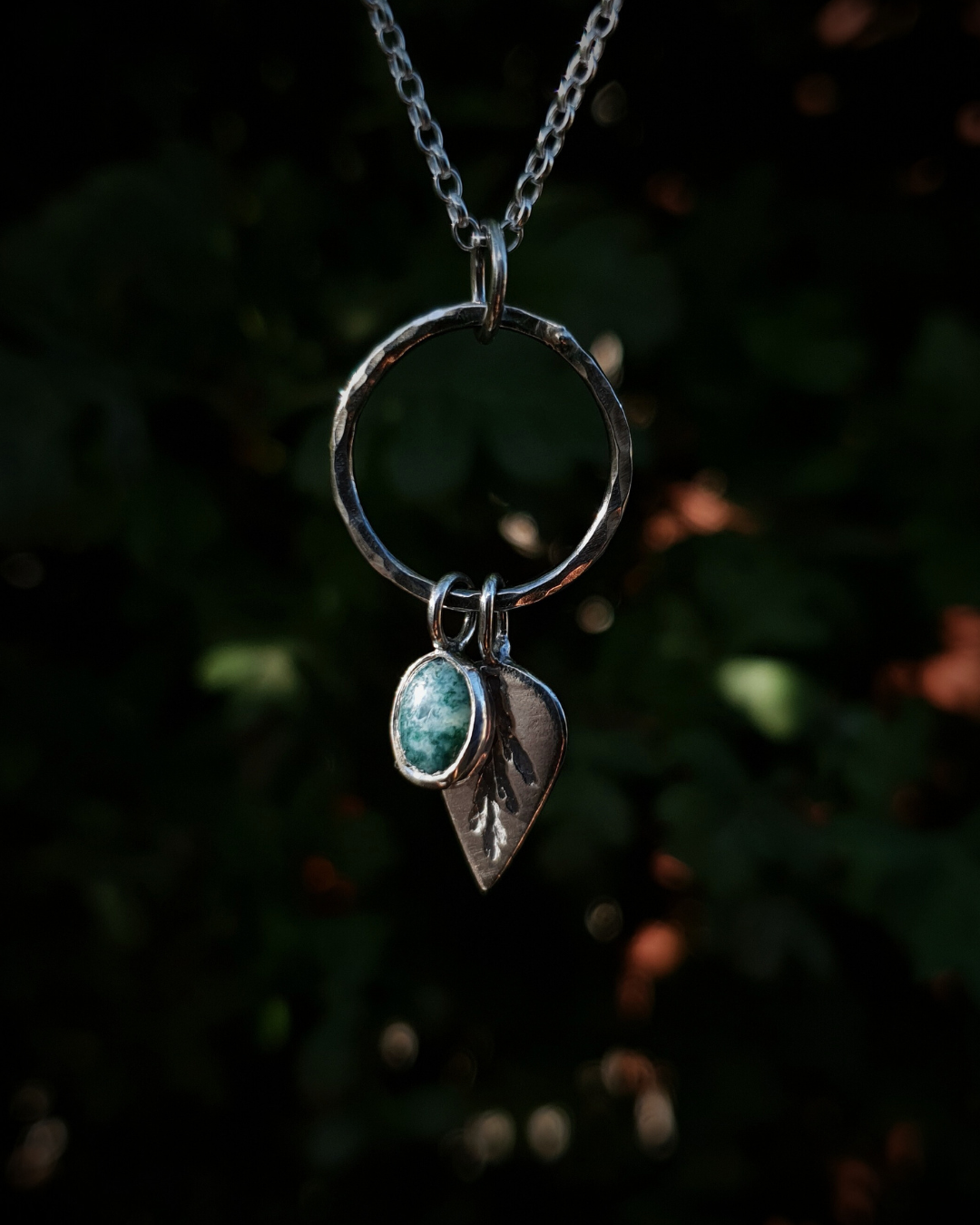 Silver pendant comprised of a silver circlet from which a solid silver upside-down teardrop, imprinted with a cypress sprig, and moss agate pendant hang from a silver chain. The moss agate is a small oval with dark green and white details; and the teardrop charm is approximately twice it's length. In the background is out of focus natural greenery.