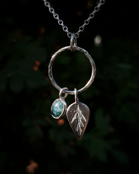Silver pendant comprised of a silver circlet from which a solid silver upside-down teardrop, imprinted with a cypress sprig, and moss agate pendant hang from a silver chain. The moss agate is a small oval with dark green and white details; and the teardrop charm is approximately twice it's length. In the background is out of focus natural greenery.