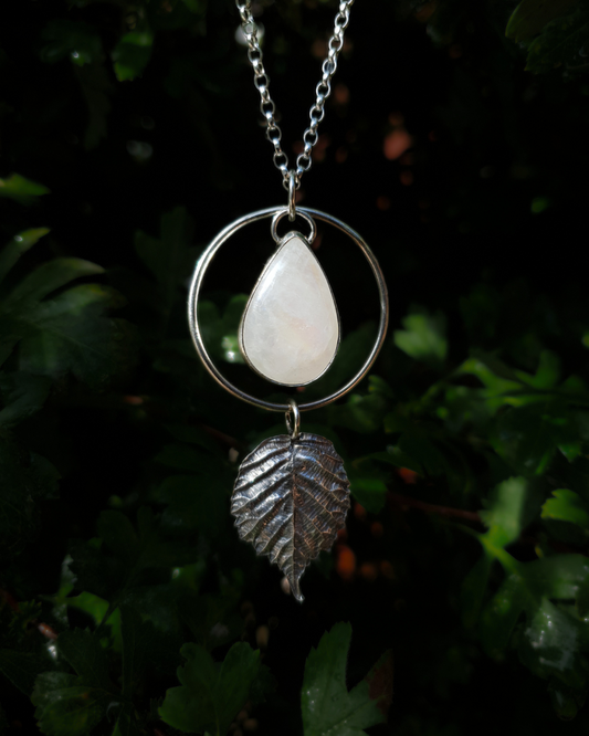 A silver pendant hangs from a chain in front of dark green, out-of-focus background. It is comprised of a large, smooth, teardrop-shaped rainbow moonstone bezel-set in silver and surrounded by a hammered silver halo-like circlet. A solid silver bramble leaf hangs rom the bottom of the circlet. Everything is attached by tiny jump rings so it can move naturally. The leaf is oxidised and polished to reveal the natural details including veins. The pendant is illuminated in natural sunlight.