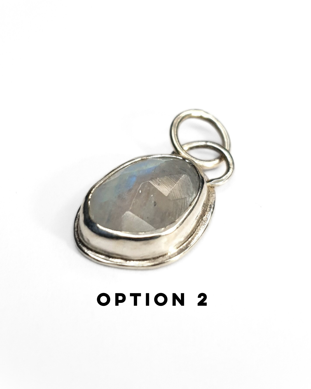 An irregular, oval-shaped rainbow moonstone is bezel set in silver and sits on a white background. The gemstone is rose-cut and the facets shine in the light displaying a beautiful show of blue-ish rainbow iridescence. There is a large circular silver bail at the top with a similarly large jump ring attached. There is text below which reads "OPTION 2"