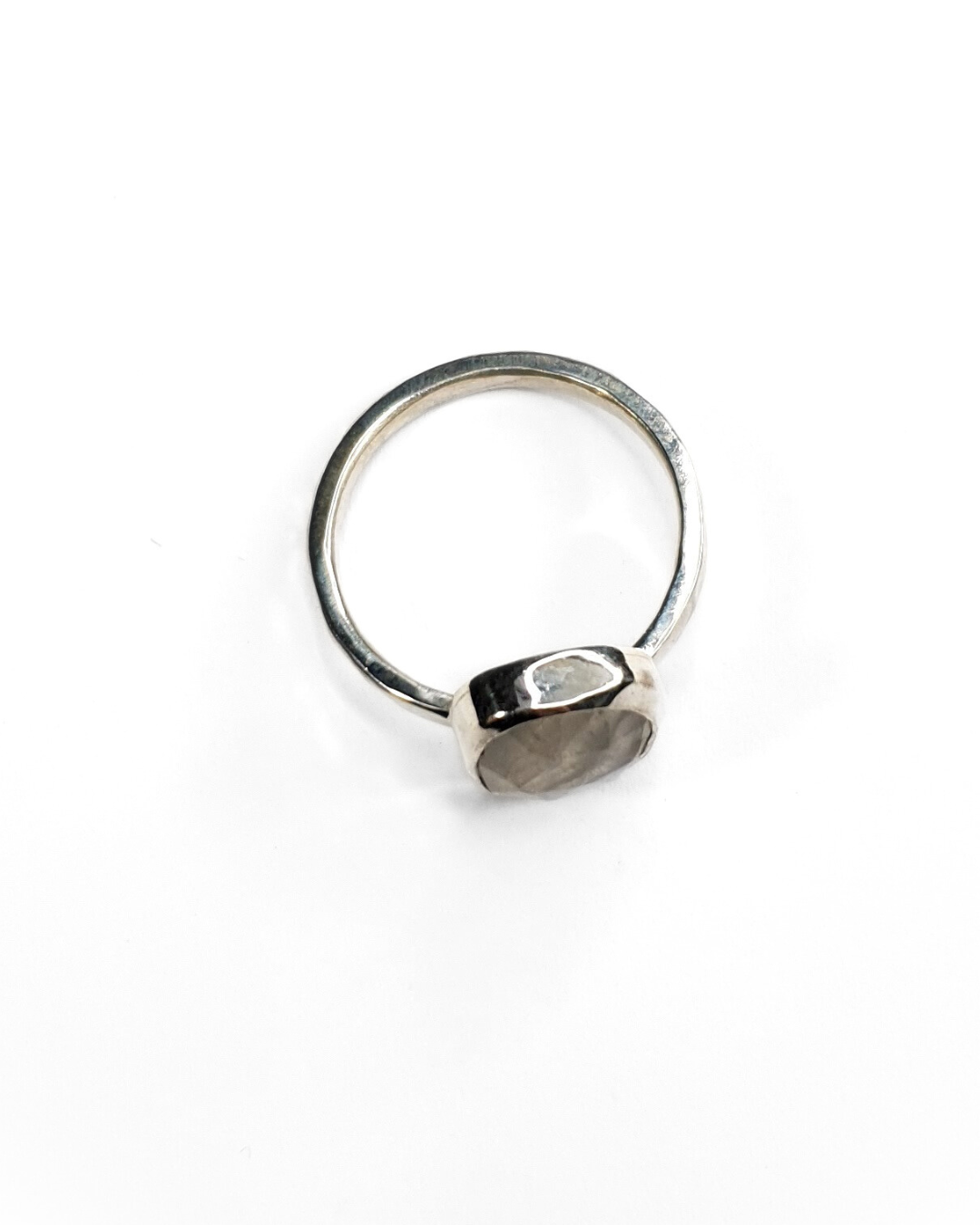 A freeform rainbow moonstone ring with a bezel setting on a shiny, hammered sterling silver band. The gemstone is faceted and contains a gorgeous iridescence and inclusions. The background of the photo is white. The photo is taken from the top with the ring laying flat on a white surface so you can see the profile.