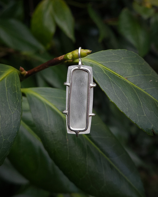 A rectangular, clear quartz pendant hangs from a thin branch, amongst large green leaves. The photograph shows the pendant from the back to demonstrate how it's set in a simple, sterling silver setting to allow the light to shine through and illuminate the stone.