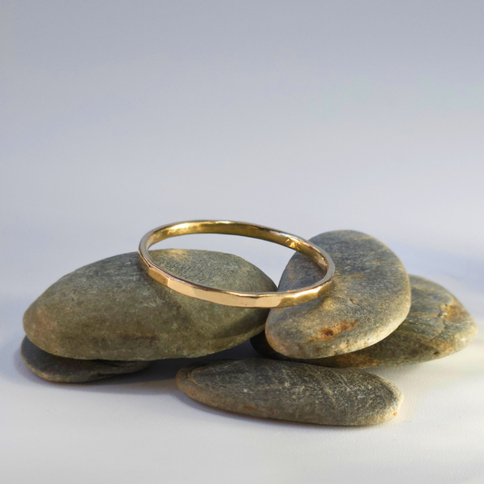 A delicate, recycled 9ct yellow gold ring sits on a small pile of pebbles on a light, neutral backdrop. The light, coming in from the right, shines gorgeously, reflecting in it's faceted hammered sides.