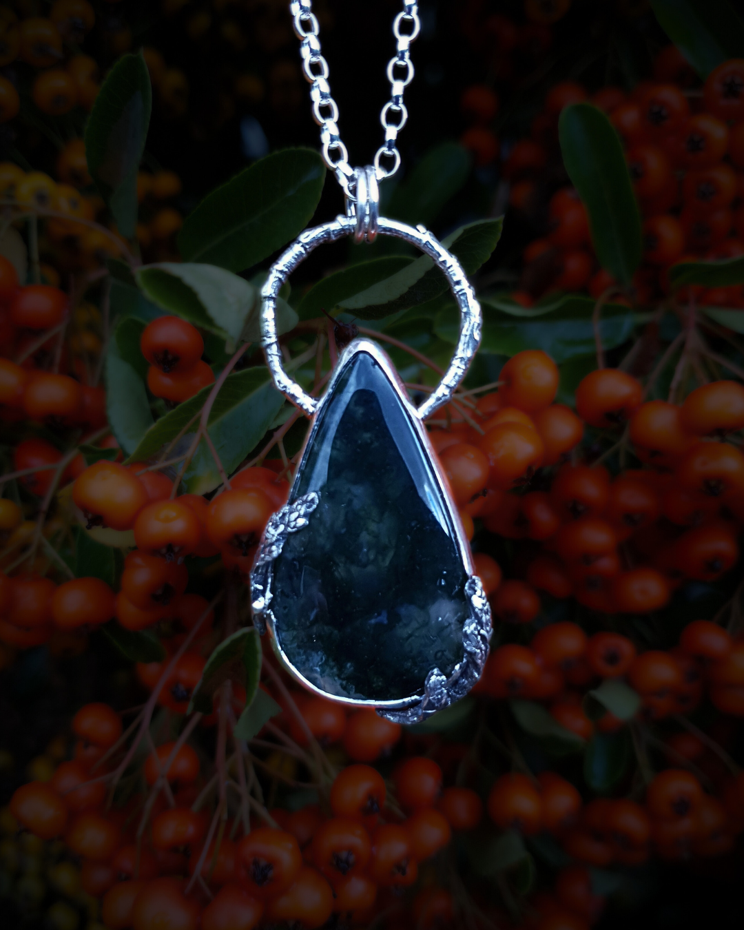 A silver pendant hangs from a chain in front of red, out-of-focus berry bush. It is comprised of a large, smooth, teardrop-shaped moss agate bezel-set in silver with a halo-like bail made from the solid-silver casting of a real twig. There are solid silver castings of cypress-sprigs surrounding the stone too. The silver is oxidised and polished back to reveal all the rich, natural textures of the twigs. The pendant is illuminated in natural sunlight showing the beautiful inclusions of the moss agate.