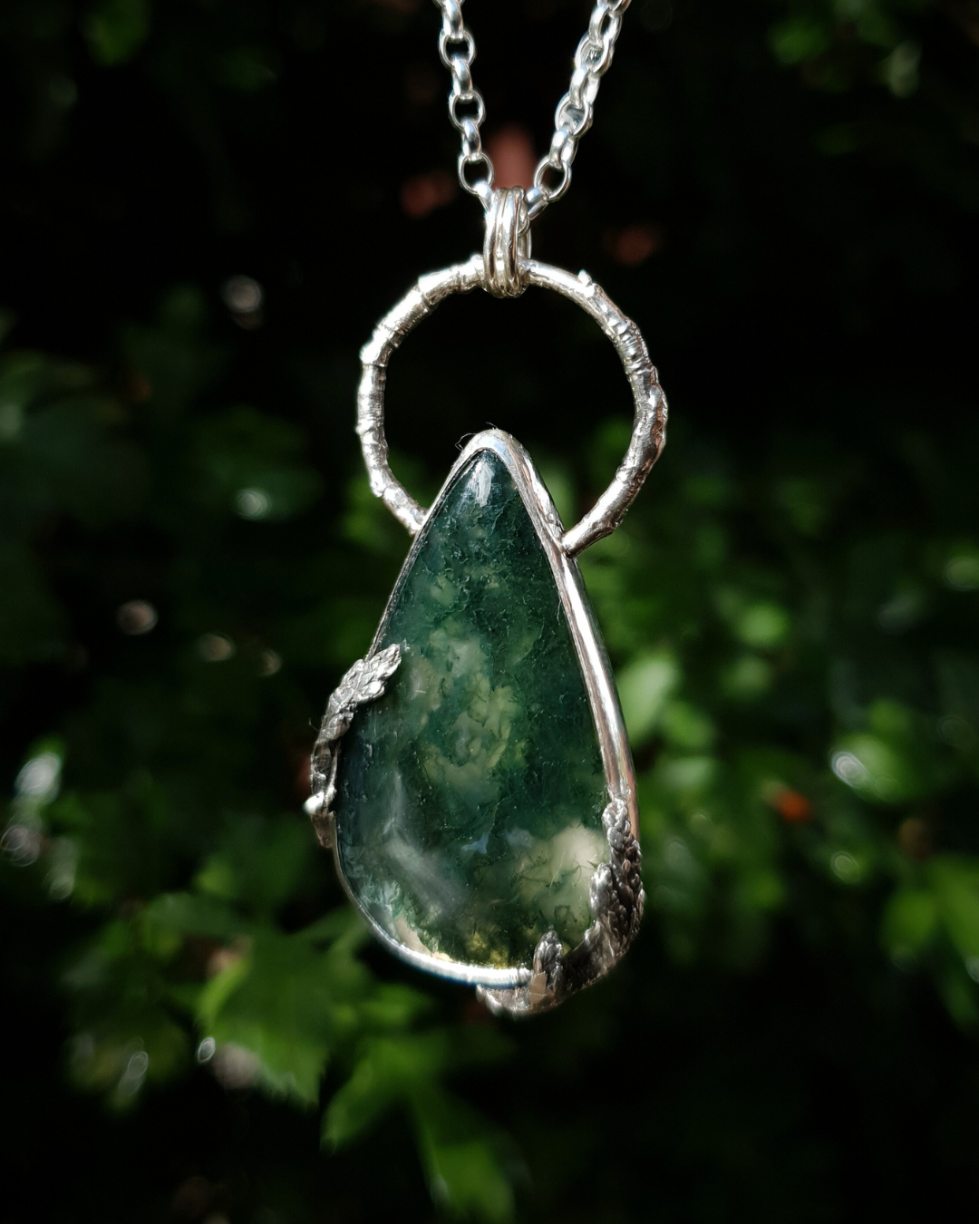 A silver pendant hangs from a chain in front of green, out-of-focus background. It is comprised of a large, smooth, teardrop-shaped moss agate bezel-set in silver with a halo-like bail made from the solid-silver casting of a real twig. There are solid silver castings of cypress-sprigs surrounding the stone too. The silver is oxidised and polished back to reveal all the rich, natural textures of the twigs. The pendant is illuminated in natural sunlight showing the beautiful inclusions of the moss agate.