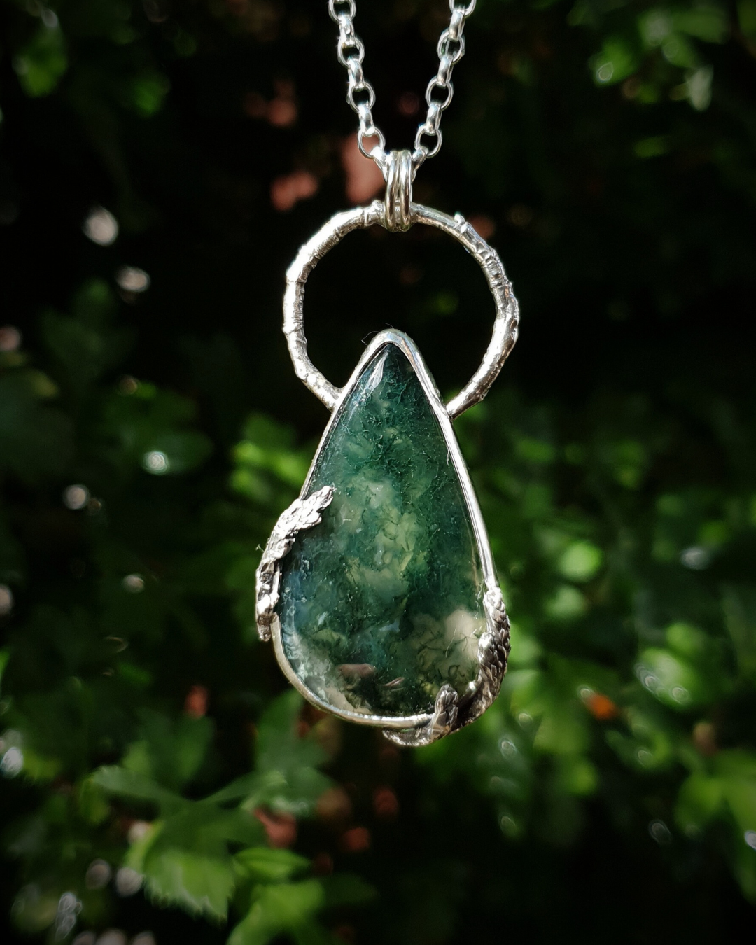 A silver pendant hangs from a chain in front of green, out-of-focus background. It is comprised of a large, smooth, teardrop-shaped moss agate bezel-set in silver with a halo-like bail made from the solid-silver casting of a real twig. There are solid silver castings of cypress-sprigs surrounding the stone too. The silver is oxidised and polished back to reveal all the rich, natural textures of the twigs. The pendant is illuminated in natural sunlight showing the beautiful inclusions of the moss agate.