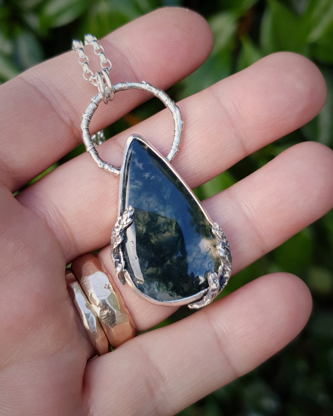 A silver pendant lays on an open palm to demonstrate its size. It is comprised of a large, smooth, teardrop-shaped moss agate bezel-set in silver with a halo-like bail made from the solid-silver casting of a real twig. There are solid silver castings of cypress-sprigs surrounding the stone too. The silver is oxidised and polished back to reveal all the rich, natural textures of the twigs. The pendant is illuminated in natural sunlight showing the beautiful inclusions of the moss agate.
