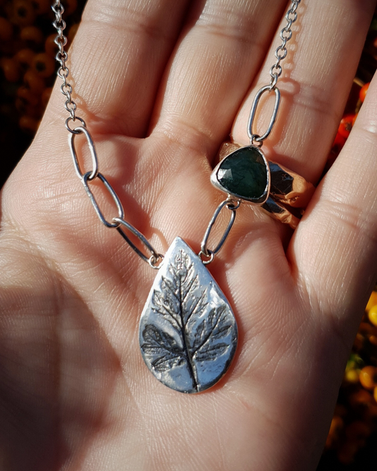 A silver necklace sits on an open palm bathed in natural sunlight. The necklace is comprised of a large silver teardrop with the oxidised impression of a leaf in it reminiscent of a fossil. To the left of the pendant there are three large oval links which join to the rest of the chain which has much smaller links. To the right of the pendant there is one large oval link, then a triangular green moss agate, bezel set in silver, and then a third large oval link which joins to the rest of the chain.