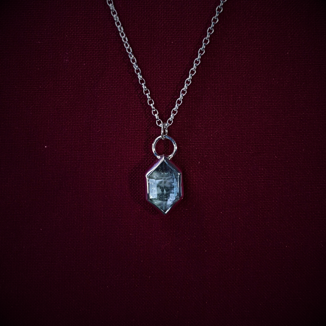 A hexagonal, .925 silver necklace hands against a burgundy backdrop. The pendant contains ashes/cremains preserved behind a faceted quartz gemstone. The ashes are visible through the gemstone which refracts the light beautifully.