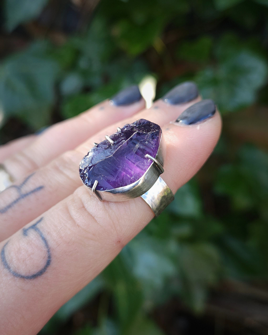 A gothic, witchy, amethyst ring sits on the end of a finger against a wall of leaves.
