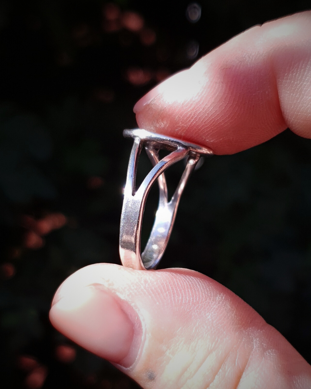 A ring is held between a finger and thumb against a dark green background to show the side profile.
The ring features a flat oval top and a split band which forks at the sides around the top.