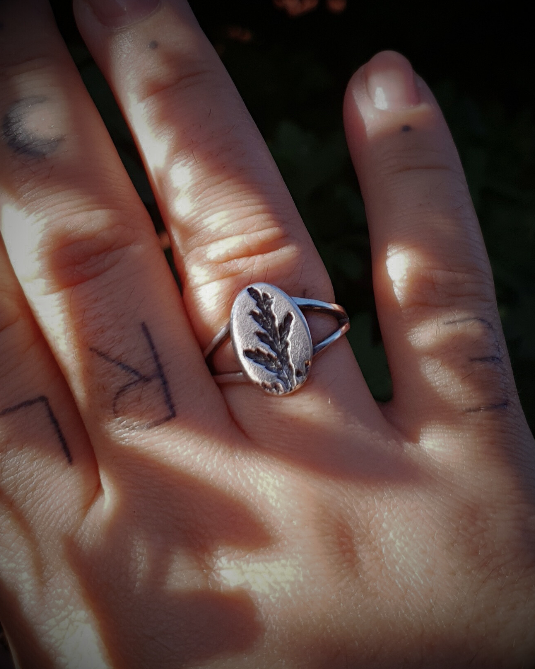 A tattooed hand wears a silver ring held up against a dark green background.
The ring features an oval top with an oxidised cypress sprig pressing and a split band.