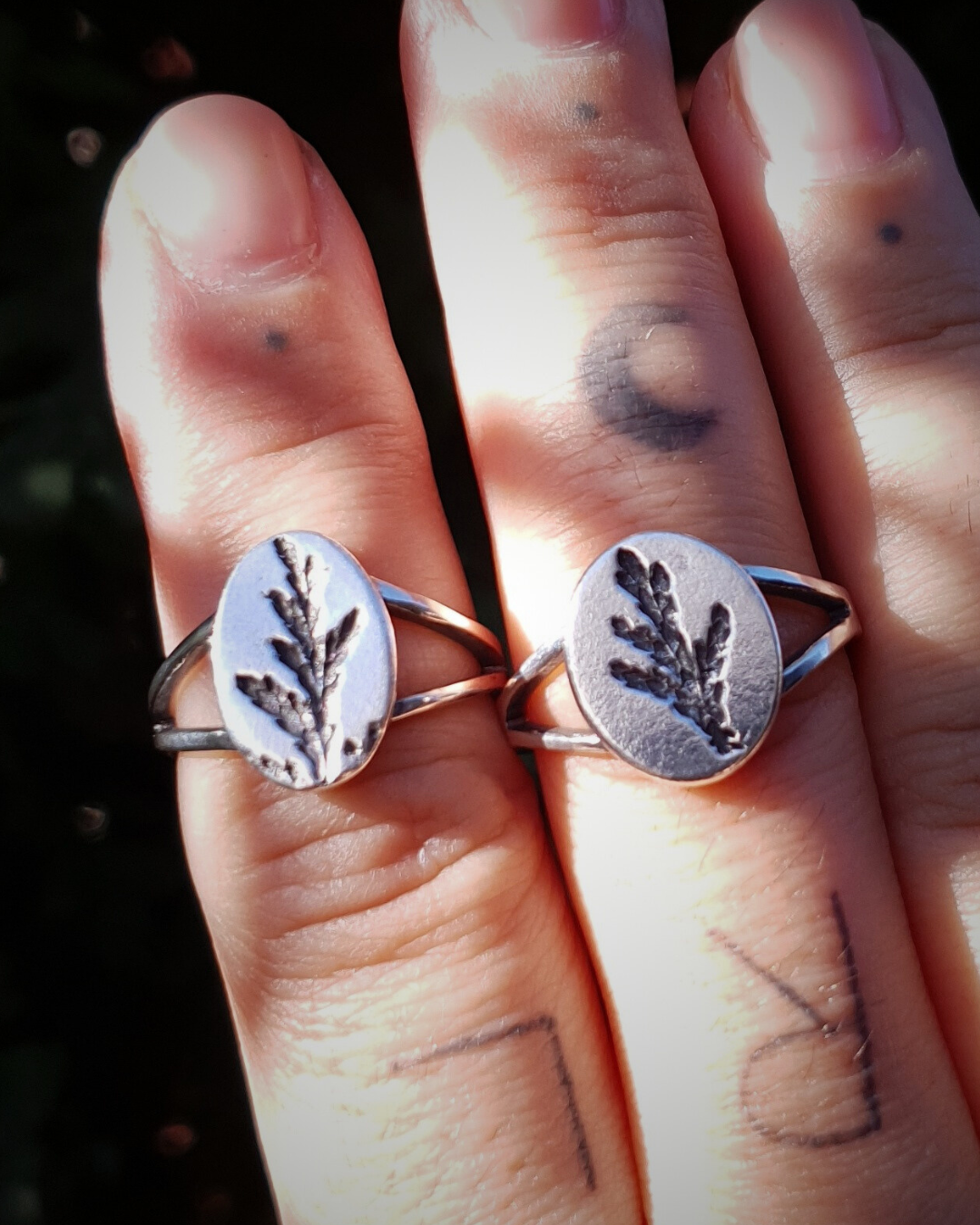 A tattooed hand wears 2 silver rings held up against a dark green background.
The rings feature an oval top with an oxidised cypress sprig pressing and a split band.