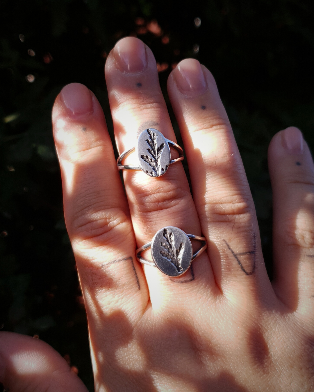 A tattooed hand wears 2 silver rings held up against a dark green background.
The rings feature an oval top with an oxidised cypress sprig pressing and a split band.