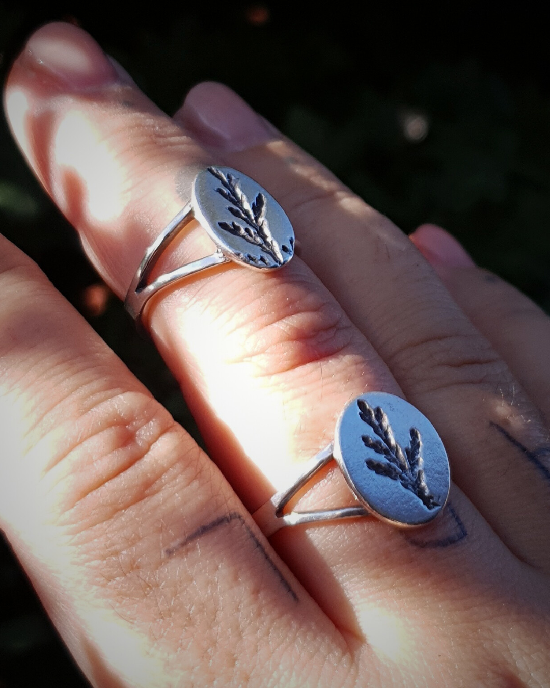 A tattooed hand wears 2 silver rings held up against a dark green background.
The rings feature an oval top with an oxidised cypress sprig pressing and a split band.