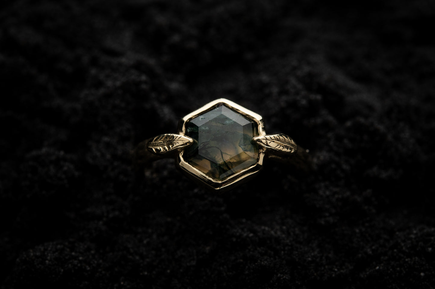A leaf-detailed engagement ring sits in black sand and is photographed from above.
A hexagonal moss agate is set in a solid gold setting with a slim leaf on either side, folded slightly over the stone.