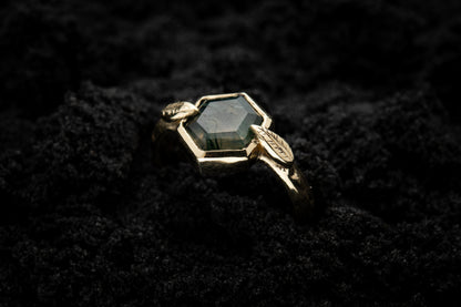 A leafy, nature inspired engagement ring is half submerged in black sand and is lit in soft, glowy light.
A lusciousy translucent, hexagonal moss agate is set in a 9 carat yellow gold setting with a slim leaf on either side, folded slightly over the stone.