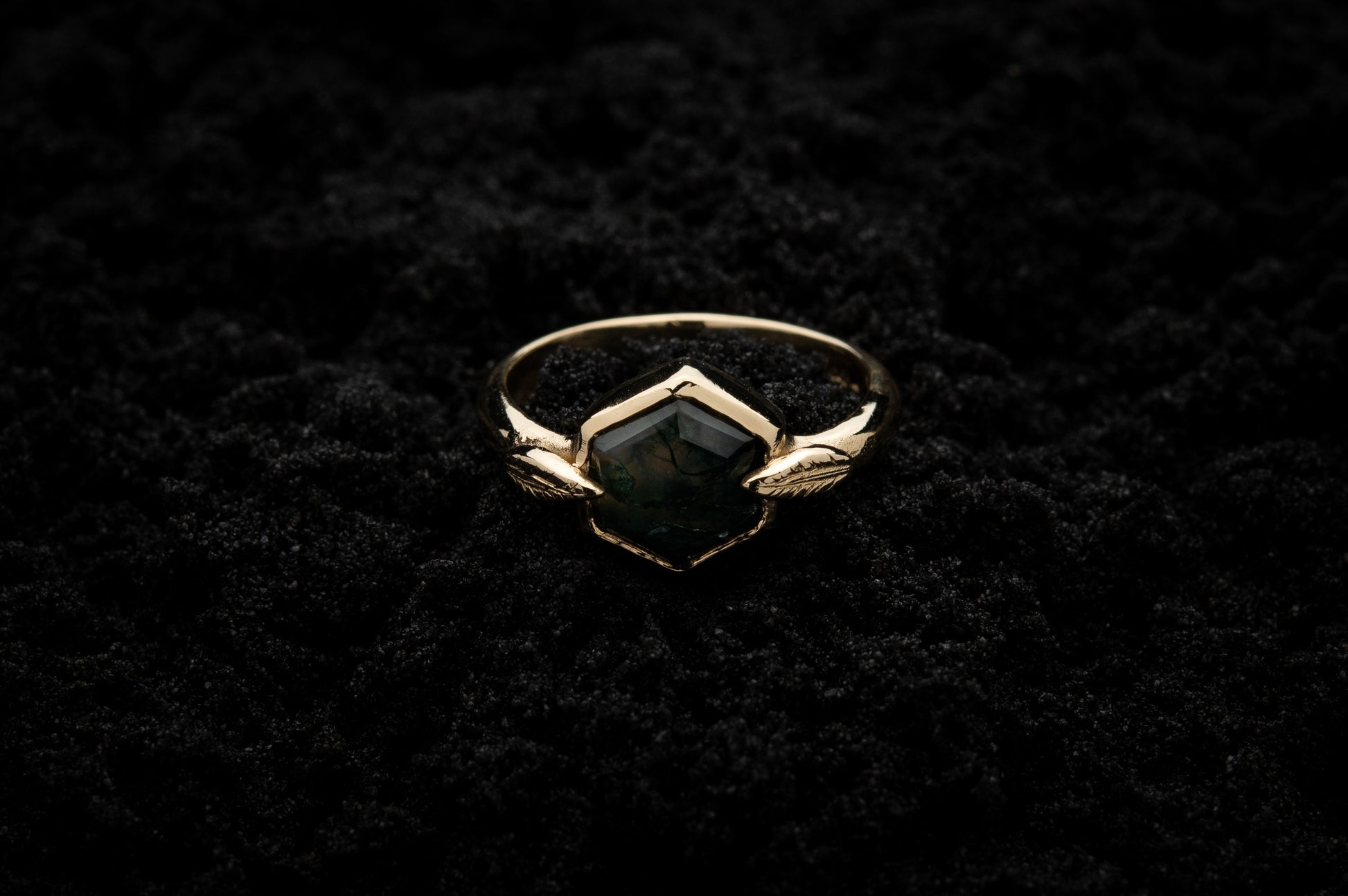 A leafy engagement ring rests on black sand and is lit in soft, glowy light.
A luscious hexagonal moss agate is set in a solid gold setting with a slim leaf on either side, folded slightly over the stone.