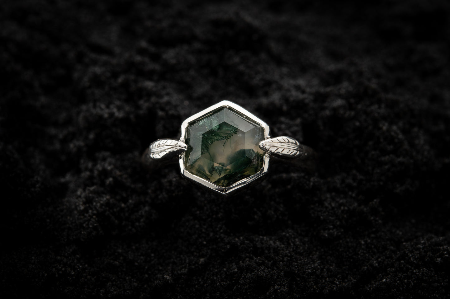 A leafy engagement ring sits in black sand and is photographed from above.
A hexagonal moss agate is set in a silvery setting with a slim leaf on either side, folded slightly over the stone.