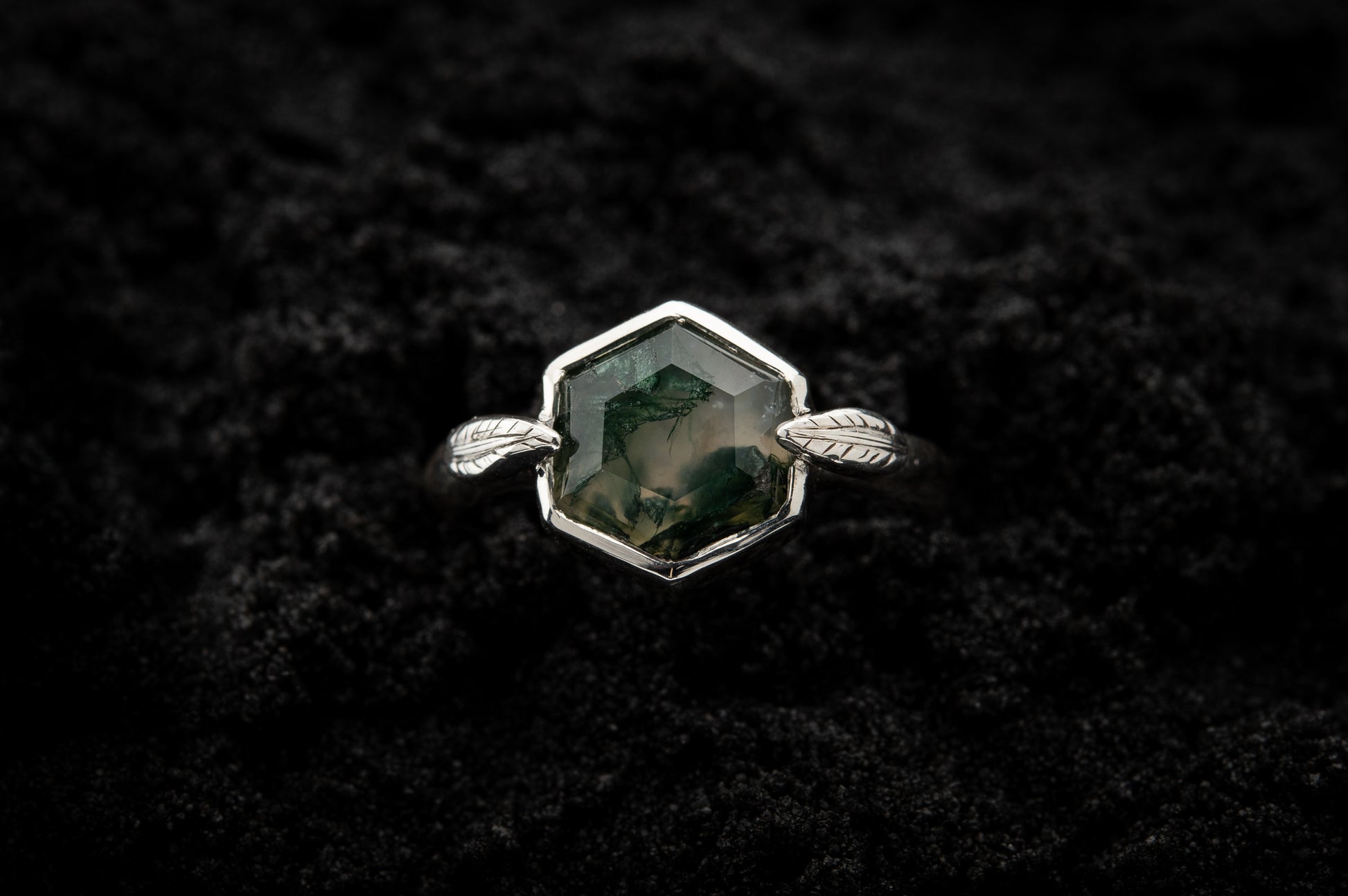 A leafy engagement ring sits in black sand and is photographed from above.
A hexagonal moss agate is set in a silvery setting with a slim leaf on either side, folded slightly over the stone.