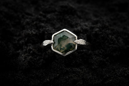 A leafy engagement ring sits in black sand and is photographed from above.
A hexagonal moss agate is set in a silvery setting with a slim leaf on either side, folded slightly over the stone.