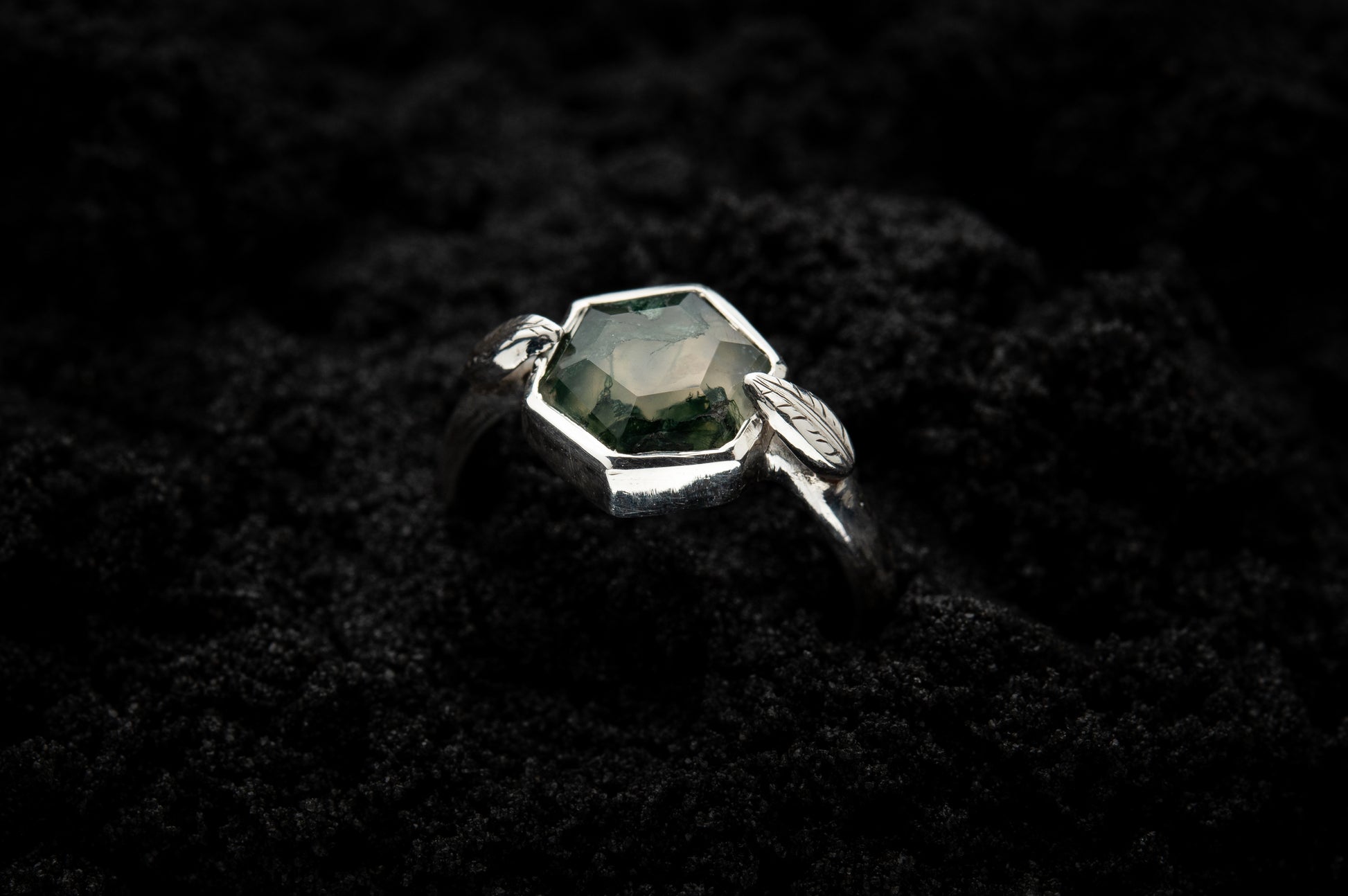 A leafy engagement ring is half submerged in black sand and is lit in soft, glowy light.
A lusciousy translucent, hexagonal moss agate is set in a white gold setting with a slim leaf on either side, folded slightly over the stone.
