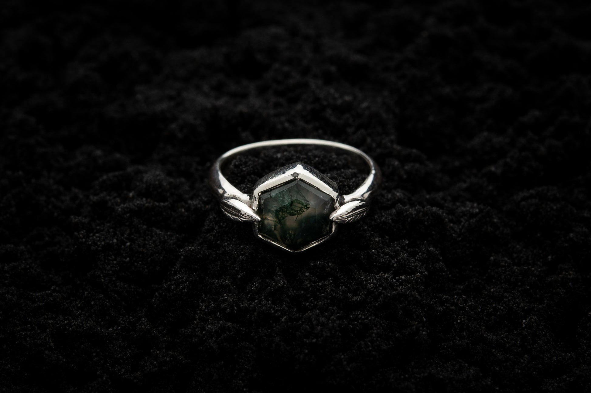 A leafy engagement ring rests on black sand and is lit in soft, glowy light.
A luscious hexagonal moss agate is set in a white gold setting with a slim leaf on either side, folded slightly over the stone.