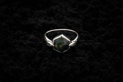 A leafy engagement ring rests on black sand and is lit in soft, glowy light.
A luscious hexagonal moss agate is set in a white gold setting with a slim leaf on either side, folded slightly over the stone.