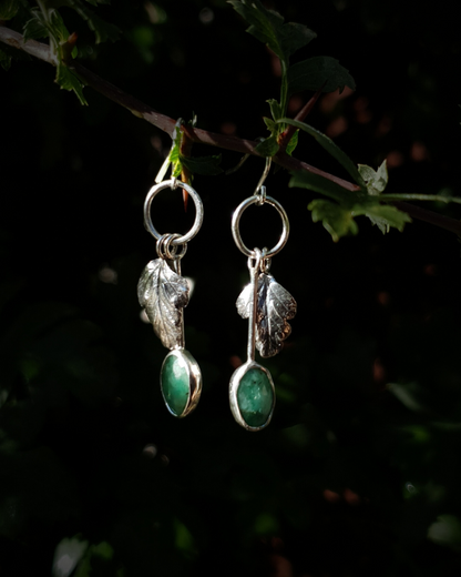 Two silver hook earrings hang from a leafy branch in front of dark green, out-of-focus background. They are comprised of a little silver circlet from which hangs a solid silver leaf. A faceted emerald also hangs from the circlet at the end of a long stem so it hangs below the leaf. The emeralds are rose-cut and rather unrefined showing different shades of green and the leaves are complete with natural details including veins. The earrings are illuminated in natural sunlight.
