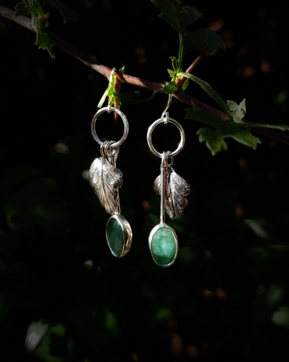 Two silver earrings hang from a leafy branch in front of dark green, out-of-focus background. They are comprised of a little silver circlet from which hangs a solid silver leaf. A faceted emerald also hangs from the circlet at the end of a long stem so it hangs below the leaf. The emeralds are rose-cut and rather unrefined showing different shades of green and the leaves are complete with natural details including veins. The earrings are illuminated in natural sunlight.