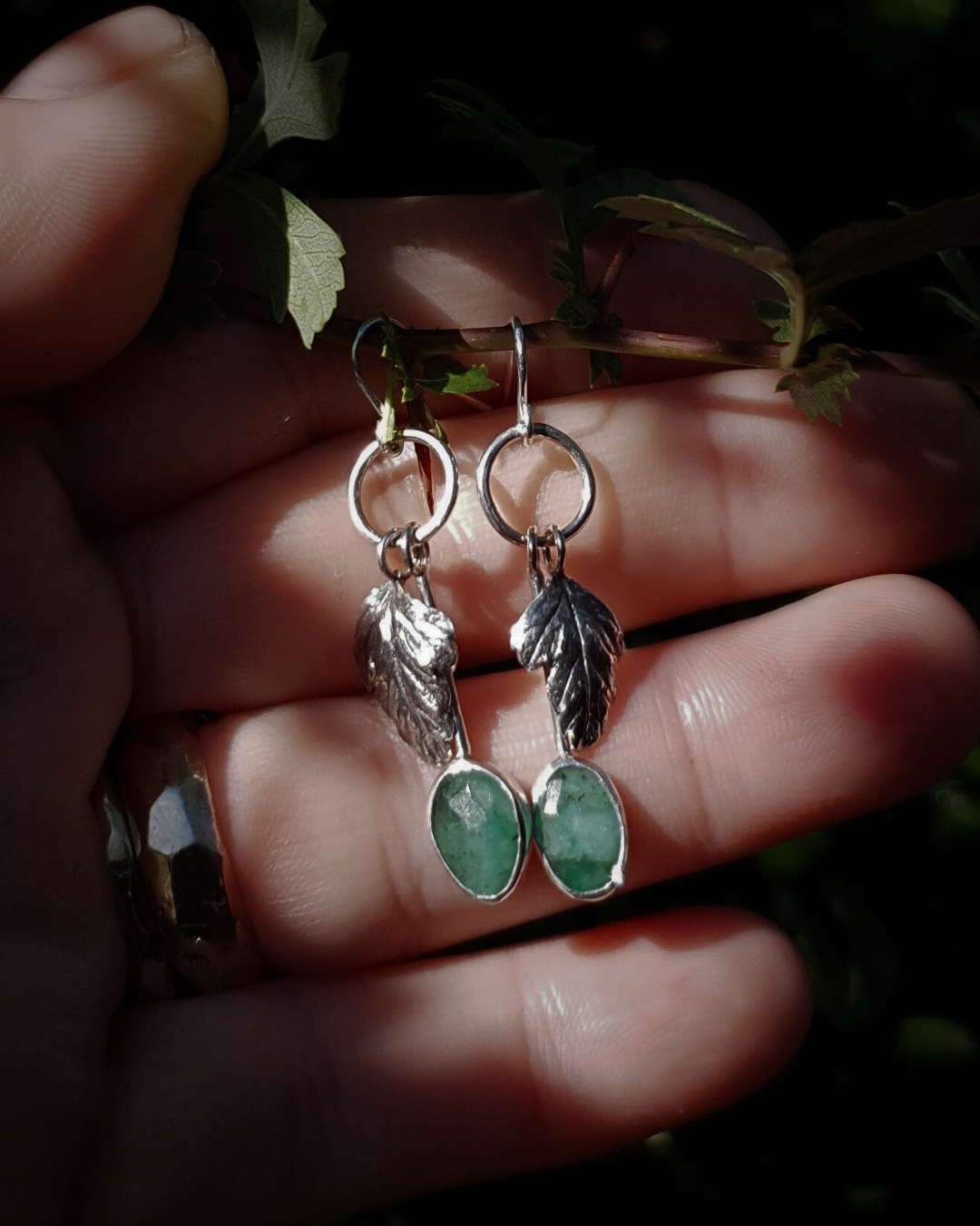 Two silver hook earrings sit in an open palm to demonstrate their size. They are comprised of a little silver circlet from which hangs a solid silver leaf. A faceted emerald also hangs from the circlet at the end of a long stem so it hangs below the leaf. The emeralds are rose-cut and rather unrefined showing different shades of green and the leaves are complete with natural details including veins. The earrings are illuminated in natural sunlight.